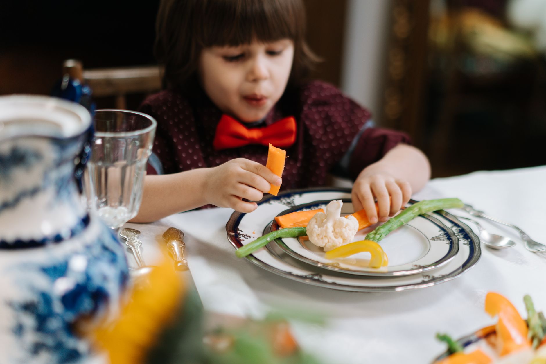 A child in a red bow tie enjoys a healthy meal with vegetables indoors.