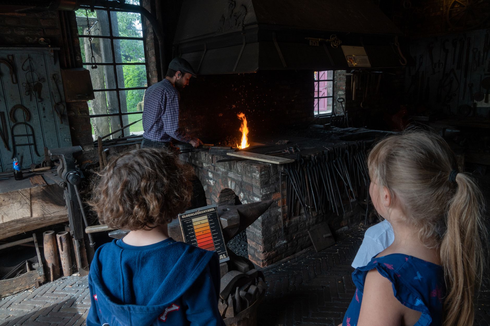 Children observe a blacksmith at work in a traditional workshop setting, showcasing cultural heritage.