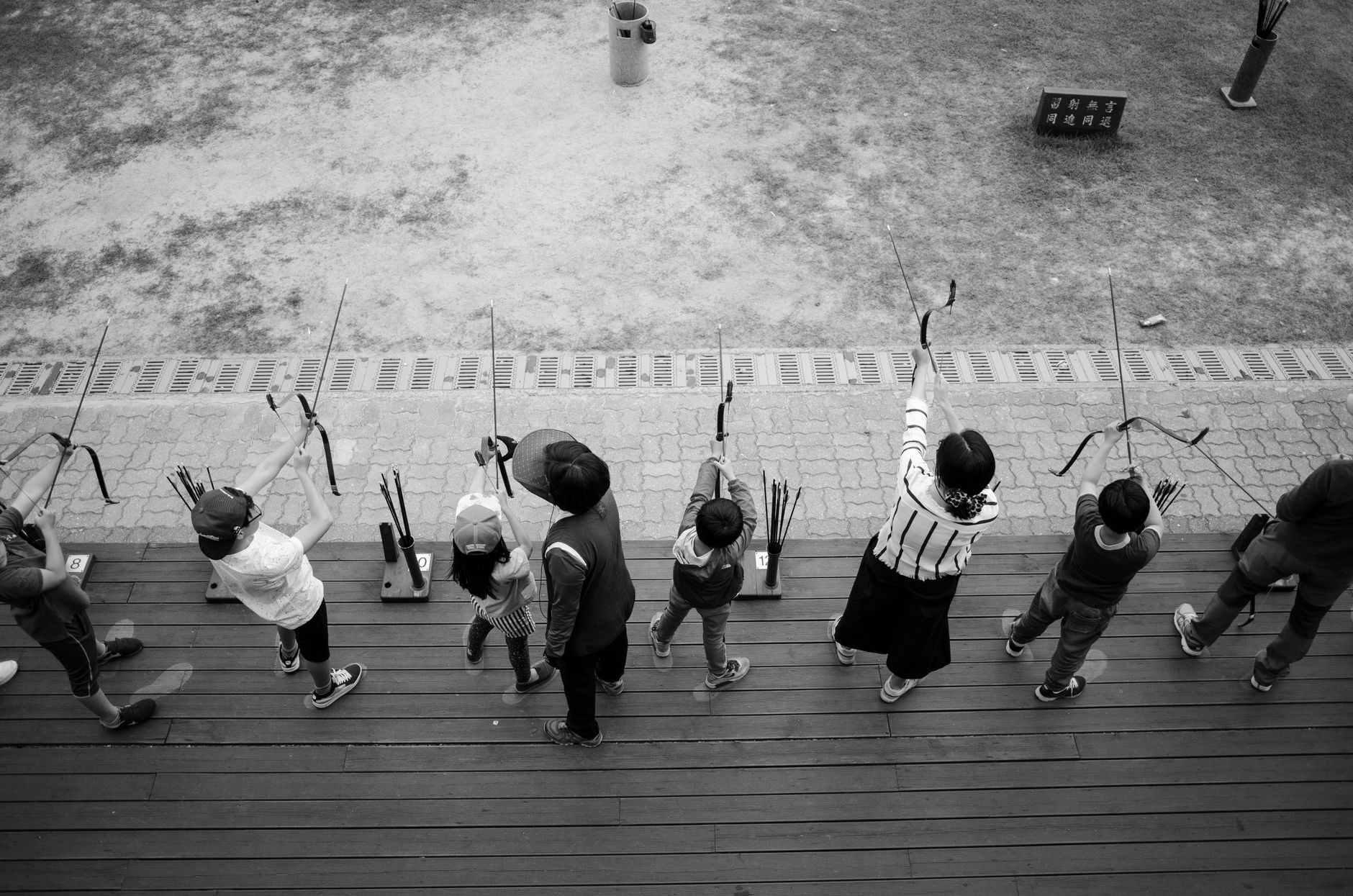 Children practicing archery outdoors, captured in a high-angle black and white shot.