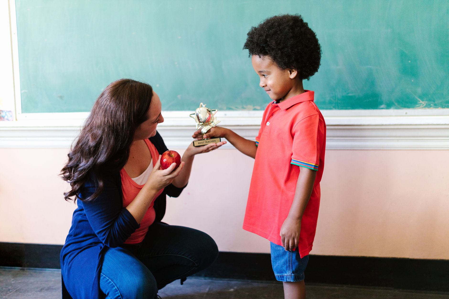 Teacher kneeling and handing a trophy to a smiling young boy in a classroom setting.