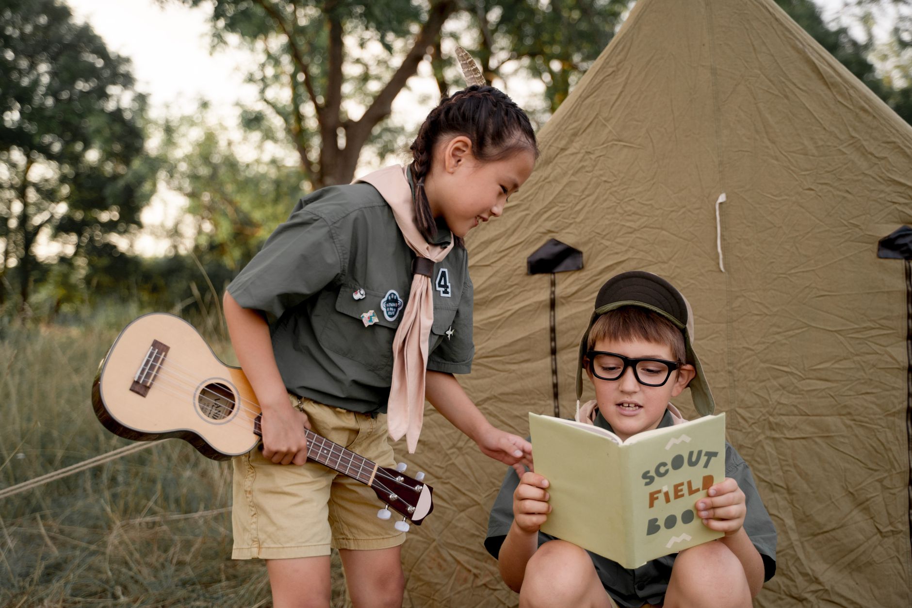 Kids in scout uniforms enjoying a camping experience with a guitar and scout field book outdoors.
