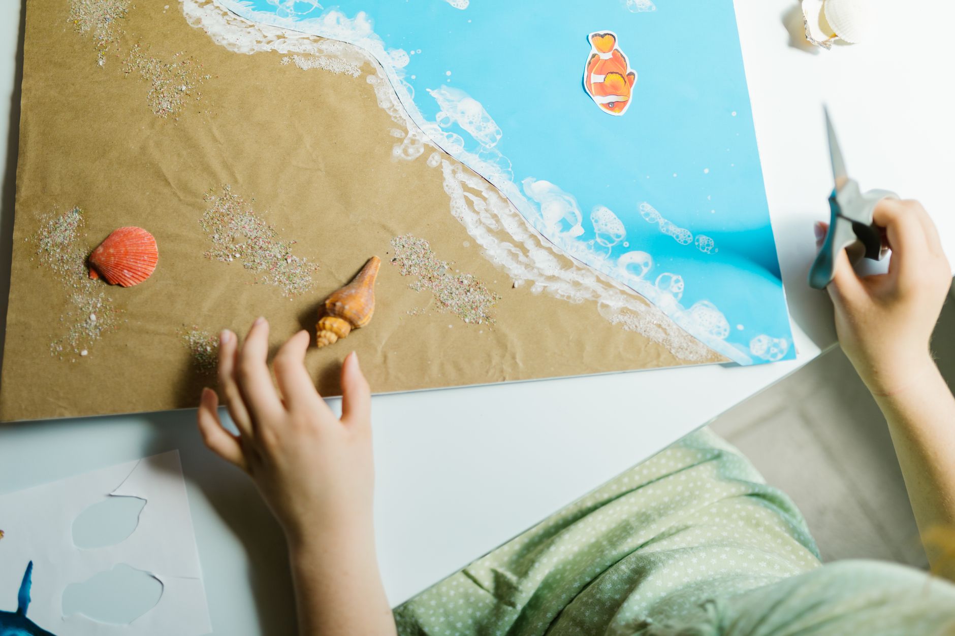 Hands crafting beach-themed collage with seashells using paper and scissors.