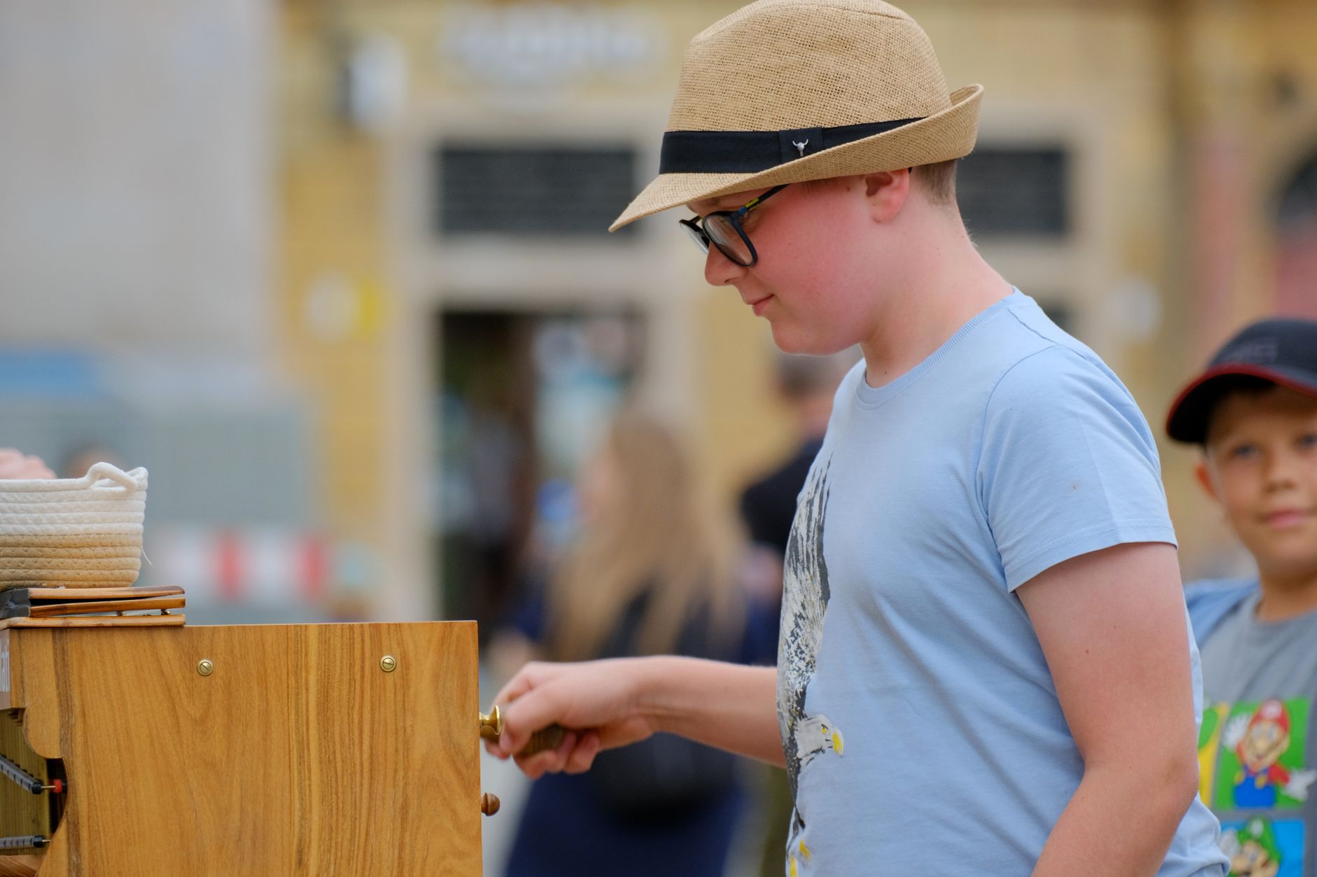 A young boy in a hat turns the handle on a street organ at an outdoor market.