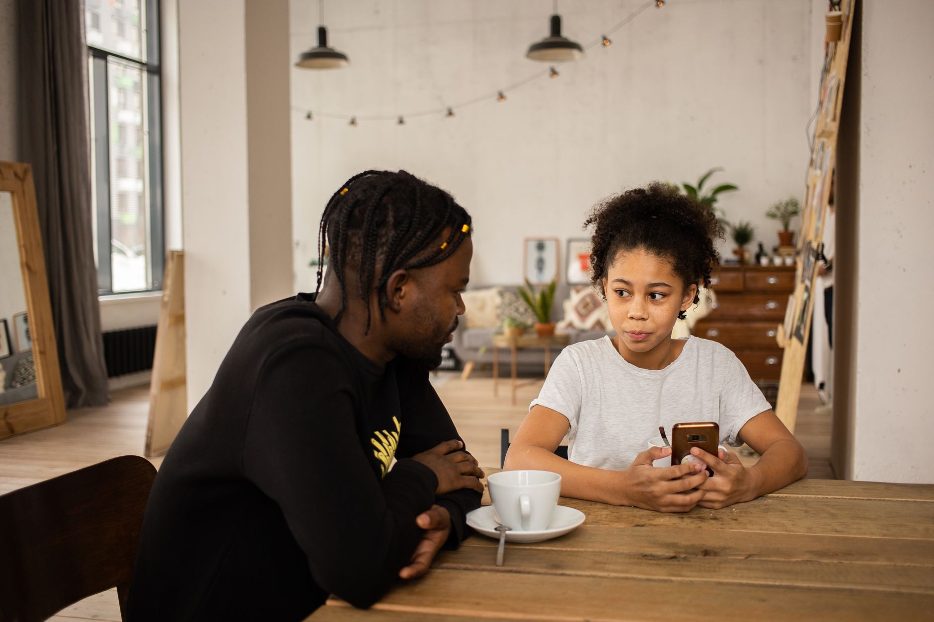 African American man sitting at table with cup of coffee and talking to daughter browsing cellphone