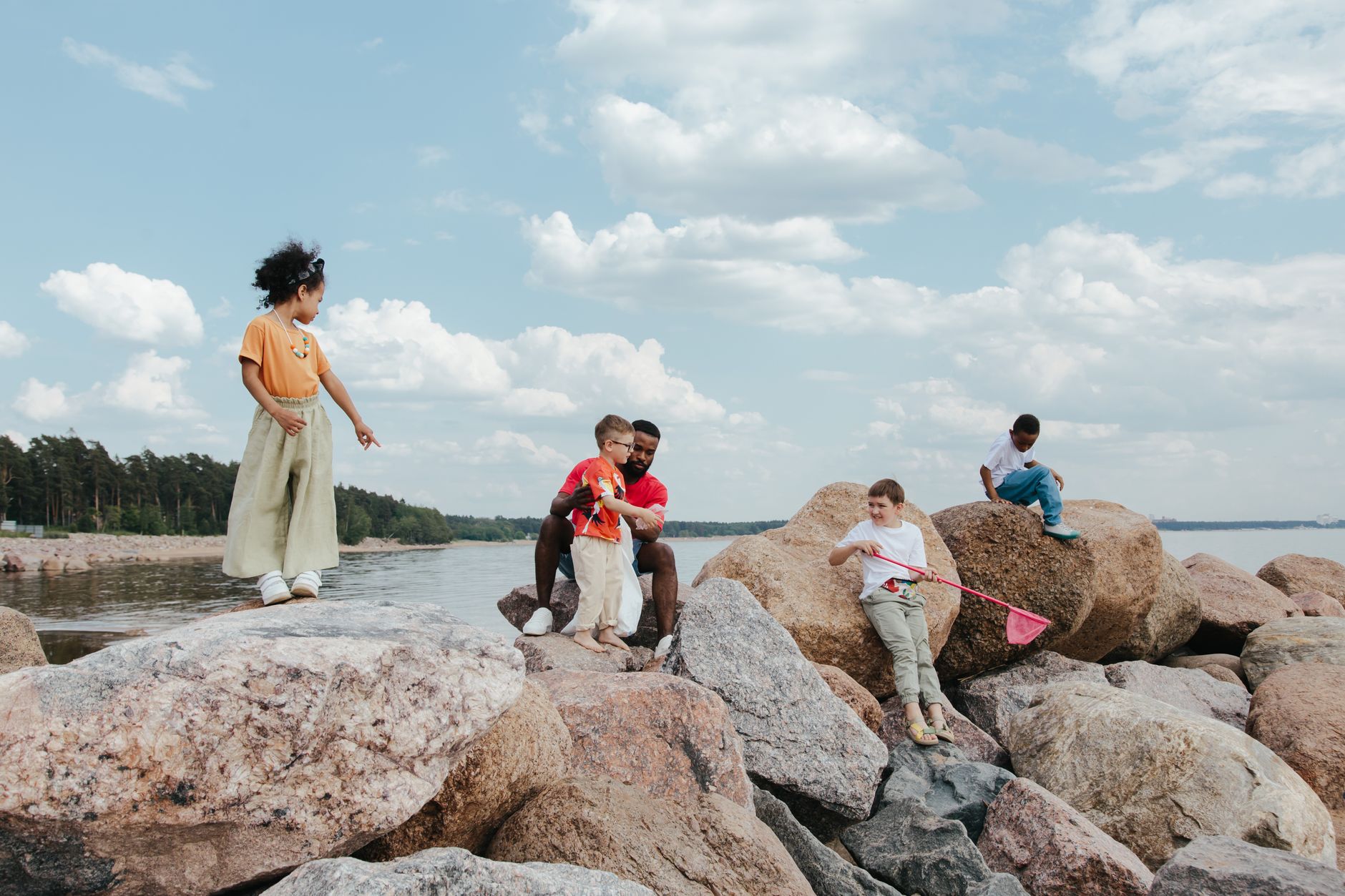 Children playing on rocks by the sea with an adult, enjoying a sunny day outdoors.