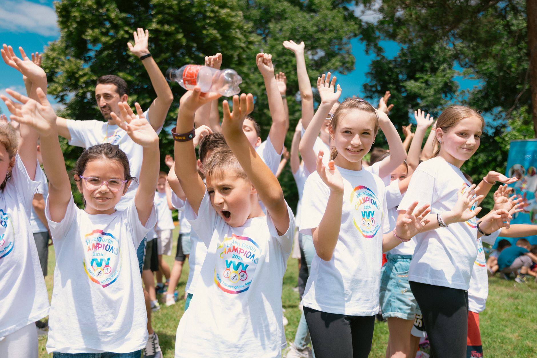 Group of cheerful children raising hands in a park during a sunny day, wearing t-shirts with event logos.