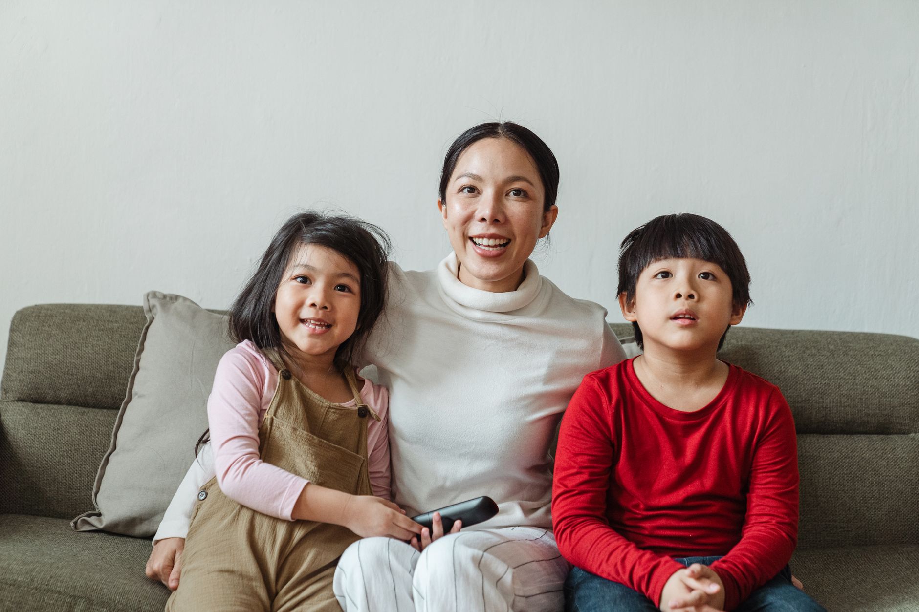 Positive Asian mother hugging little son and daughter sitting on sofa while watching funny movie on TV channel together