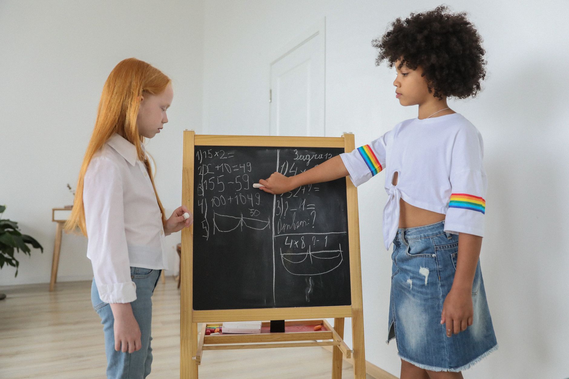 Side view of pensive girls in casual clothes leaning mathematical tasks written on easel blackboard in light room in daytime