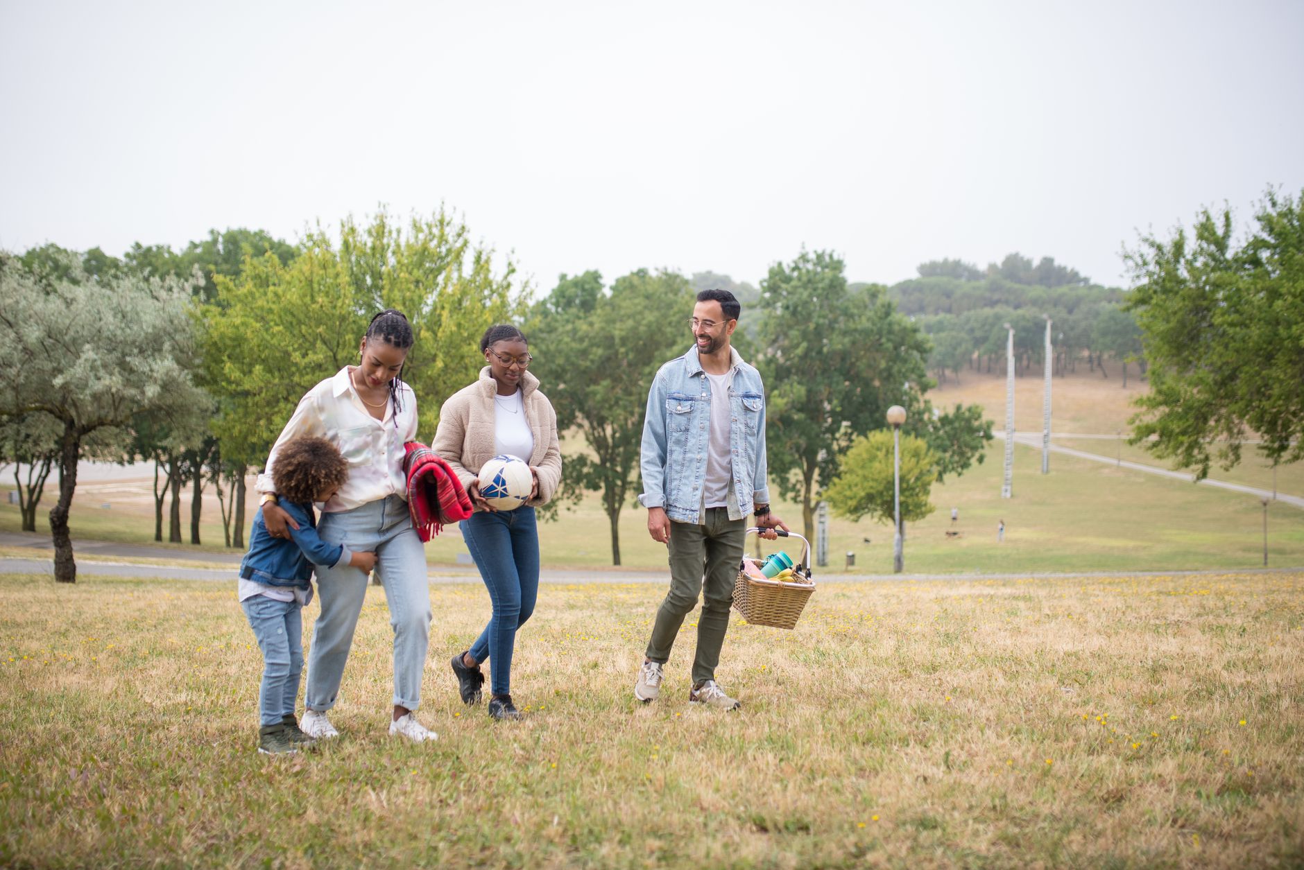 A joyful family walking through a park on a sunny day, carrying a picnic basket and a ball.