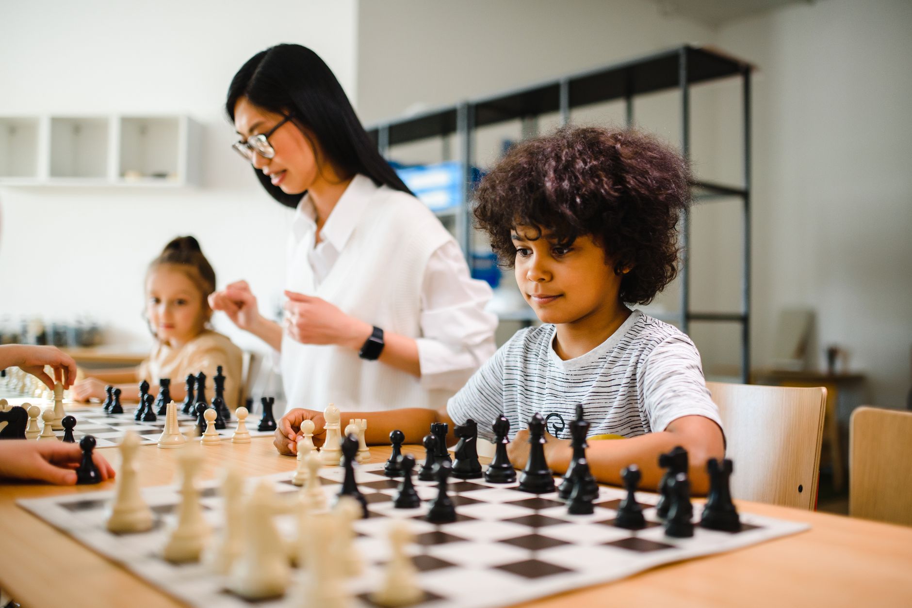 A multicultural group of children playing chess under teacher's guidance in a classroom setting.