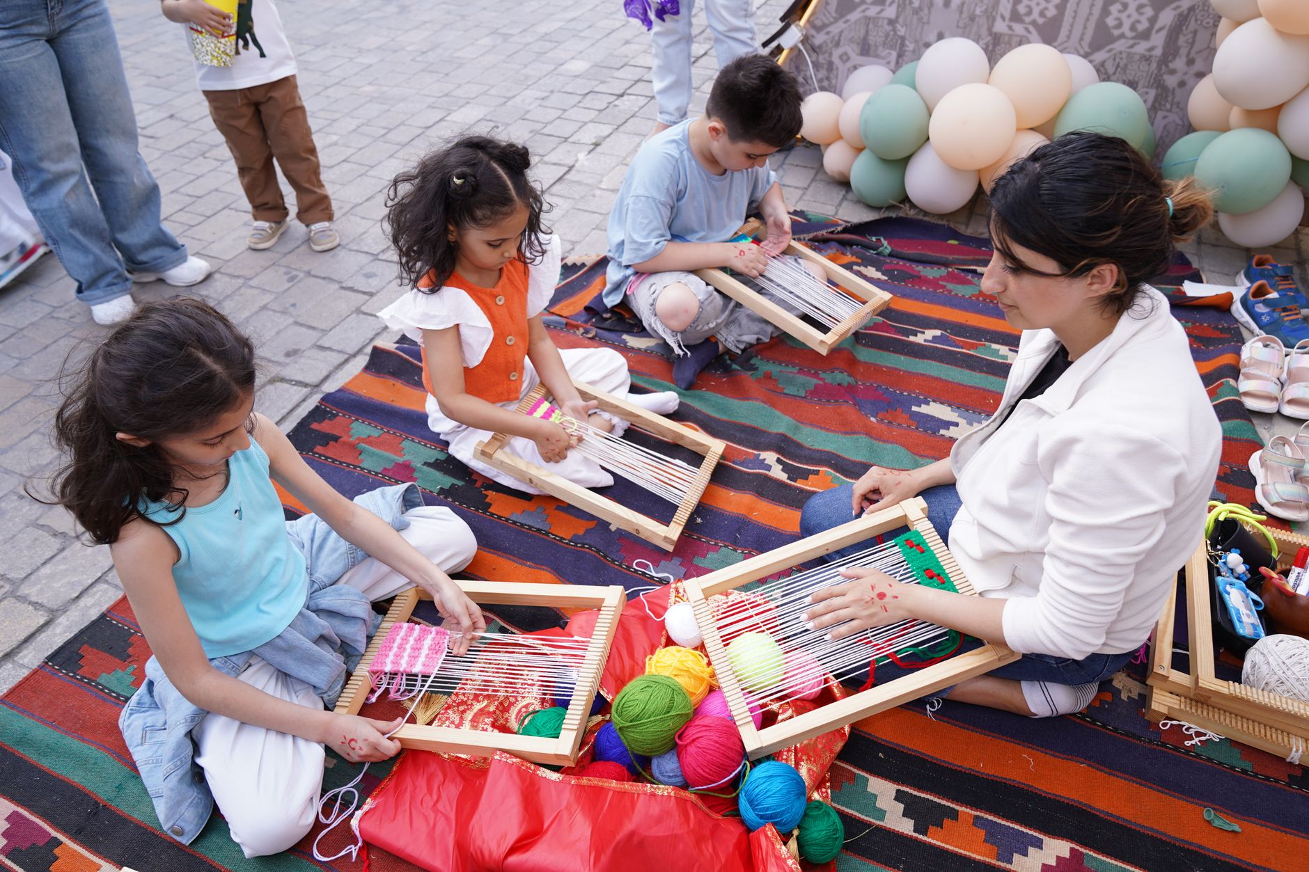 Children and a woman weaving together outdoors on a colorful mat, surrounded by yarn and balloons.