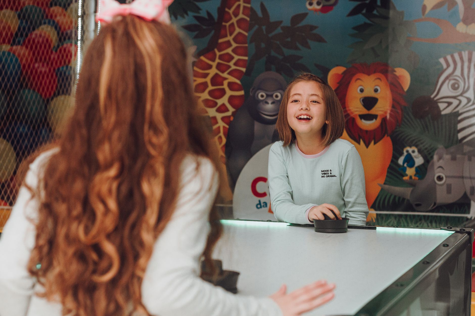 Two happy young girls playing air hockey indoors with colorful animal mural background.