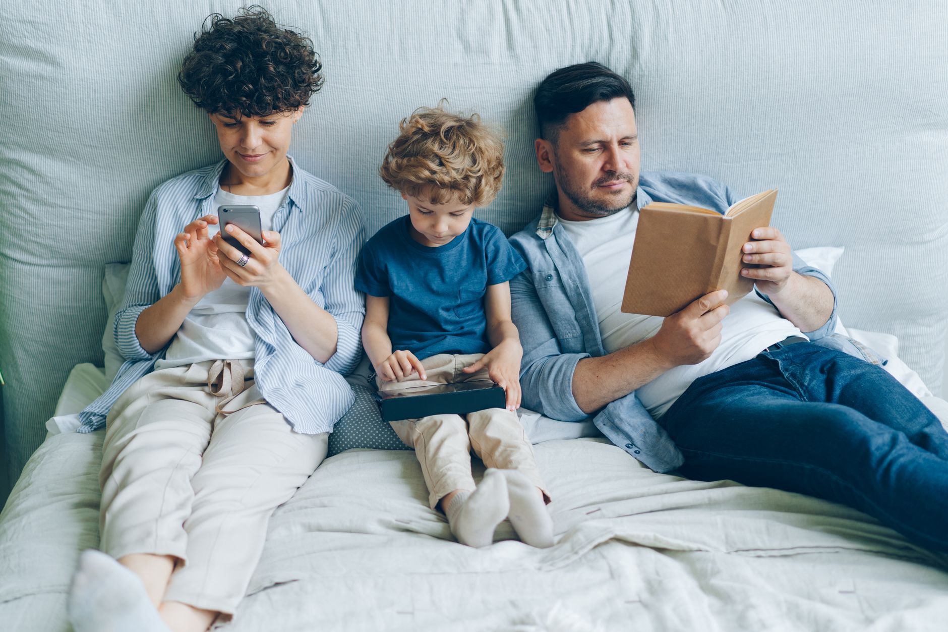 A family relaxing on bed with a smartphone, tablet, and book, symbolizing modern family life.