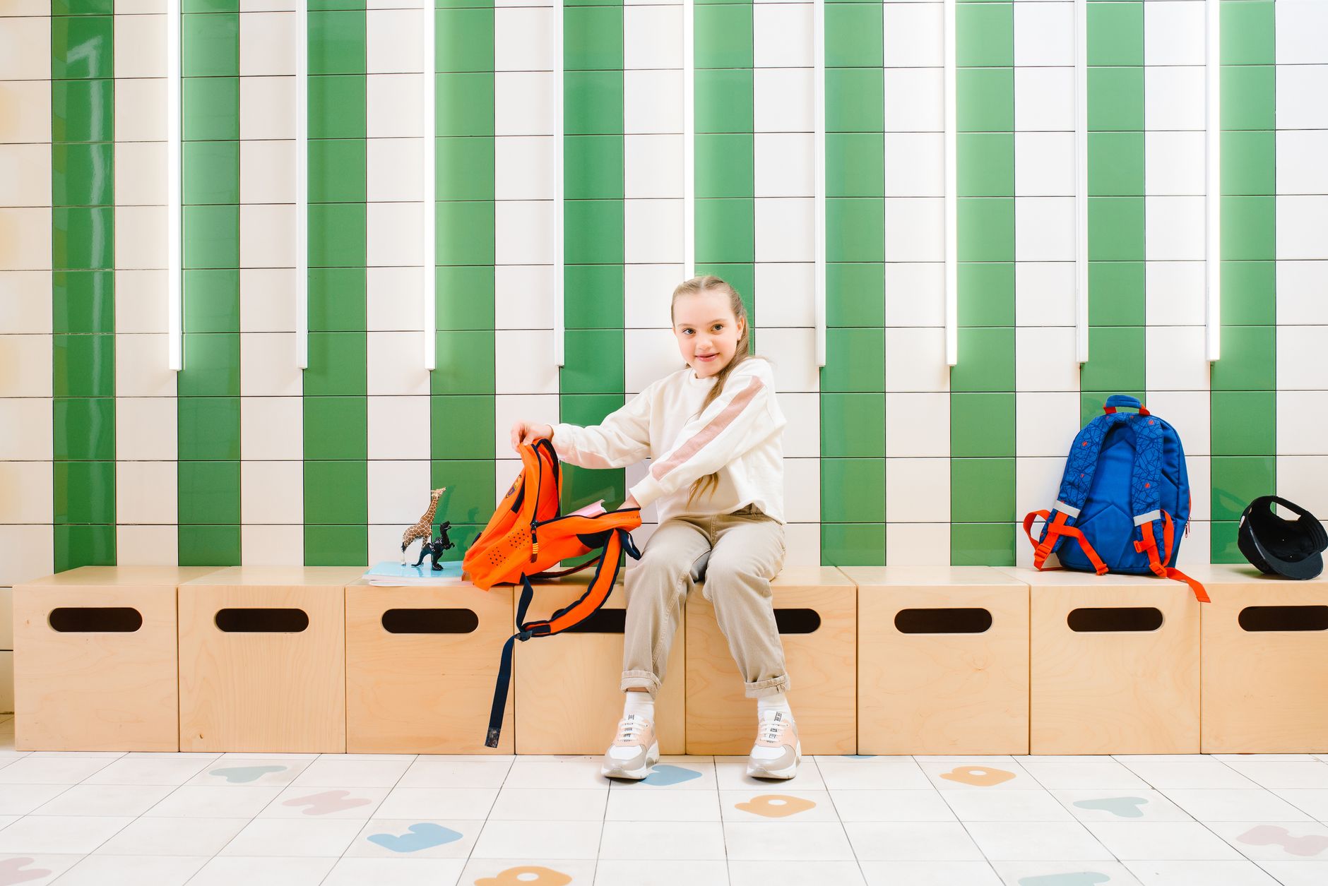 A young girl smiles while packing her backpack in a school locker room with bright decor.