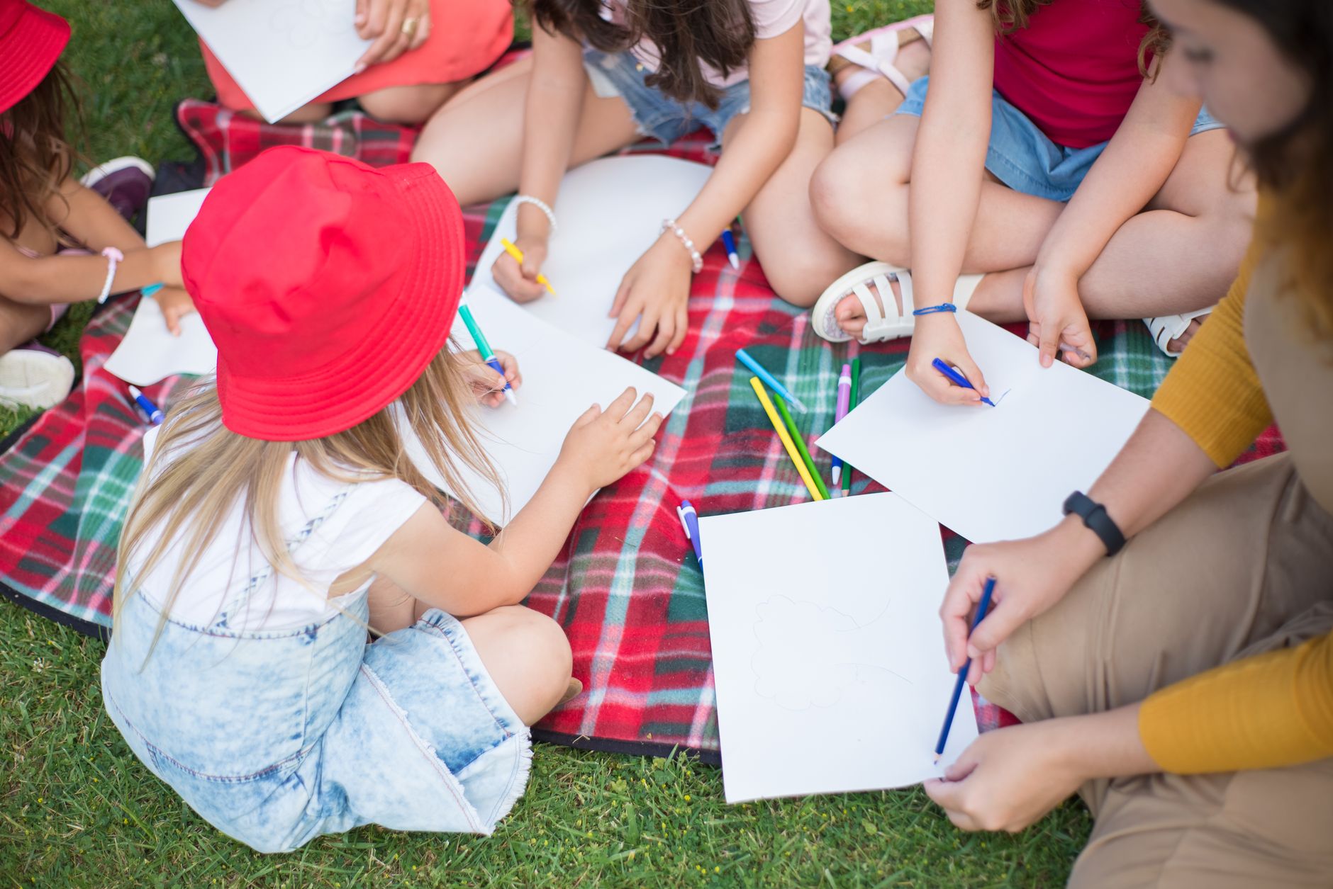 A group of children creatively drawing together on paper outdoors on a picnic blanket.