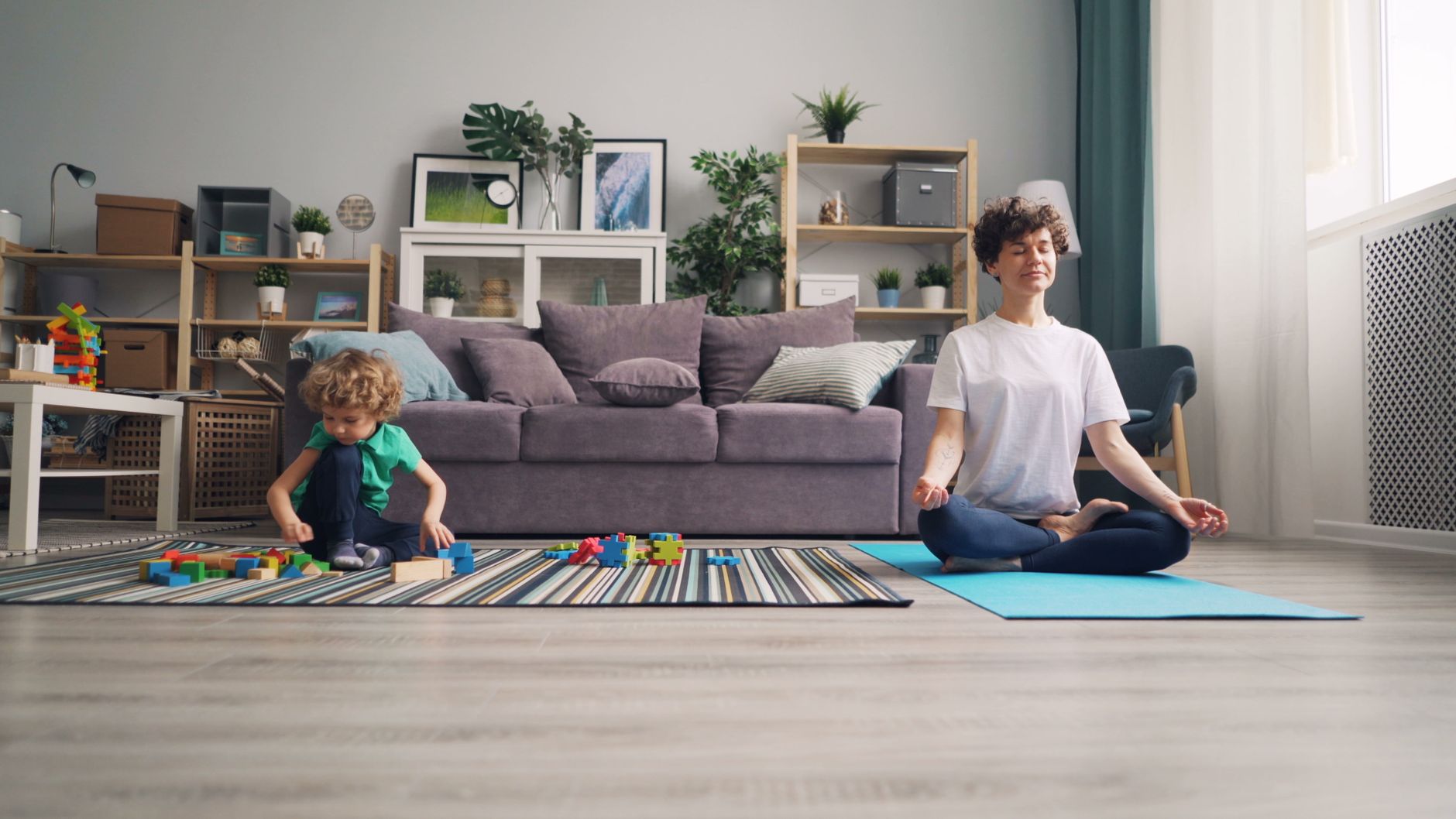 A relaxed mother meditates while her son plays with building blocks in a cozy living room.