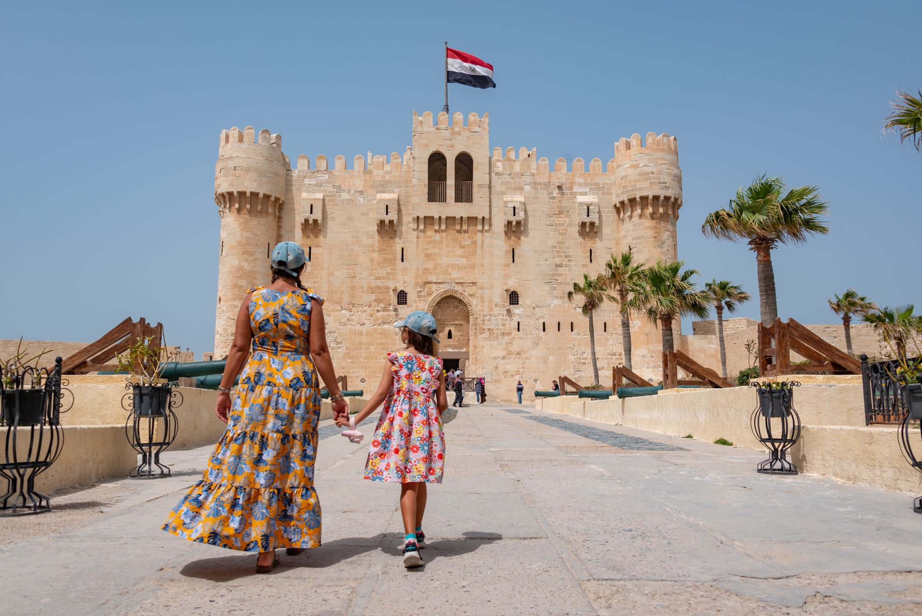 A woman and a child admire a historic fortress on a sunny day, holding hands.