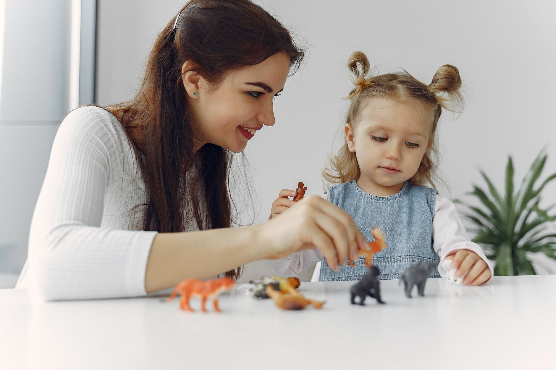 Adorable little girl sitting at white table with mother while playing with small toy wild animals for early education and development during