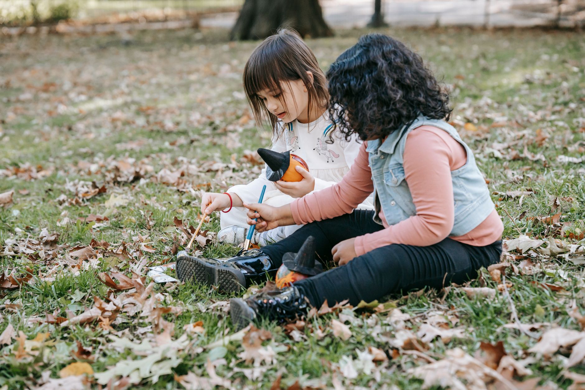 Full length of focused little girls painting on small pumpkins with brushes and gouache while sitting on grass
