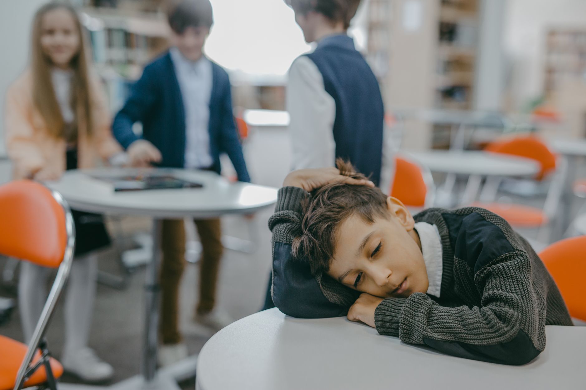 A sad child rests on a classroom desk while peers talk, symbolizing loneliness and school exclusion.